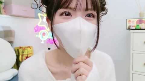 Media: Video of an Asian woman with fair skin, wearing a white mask, white top, and brown hair styled in a bun, holding a mask. Neon \"Glamour\" sign and colorful decorative items in the background.