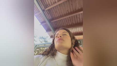 Media: A video of a young woman with wavy brown hair, wearing a white sweater, standing under a wooden-roofed structure. She appears relaxed, with her eyes closed and hand resting on her cheek.