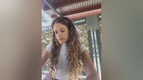 Media: Video of a young woman with long, curly brown hair, wearing a white t-shirt, standing in a sunlit outdoor space with red-tiled roof and greenery in the background.