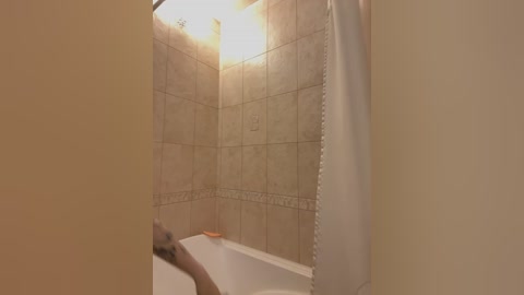 Media: Video of a beige-tiled shower with a white curtain partially drawn, revealing a white bathtub with a soap bar. The room is dimly lit, and the walls have a decorative border.