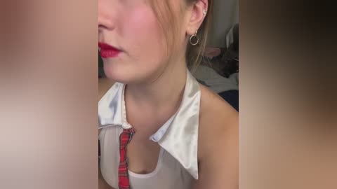 A close-up video of a young woman with fair skin, red lipstick, and a white sleeveless top with a red tie. She wears small hoop earrings and has her blonde hair tied back. The background is blurred, focusing on her face and neck.