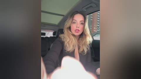 Media: Video of a Caucasian woman with long, wavy blonde hair, wearing a black jacket, sitting in a car, with a blurred view of the city outside.