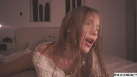 Video of a young woman with long, wet hair, leaning forward on a bed in a dimly lit room, wearing a white off-shoulder sweater.