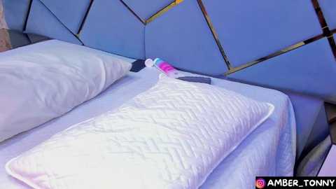 Media: Video of a modern bedroom featuring a bed with a quilted white pillow, a blue geometric-patterned headboard, and a colorful vibrator resting on the pillow.