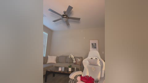 Video of a modern living room with beige walls, a gray sectional sofa, a white gaming chair, a ceiling fan, and a red item on the floor.