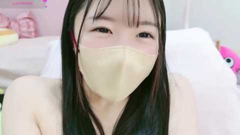 Media: Video of an Asian woman with long black hair, fair skin, wearing a beige face mask, lying on a white bed, surrounded by pink and white pillows.