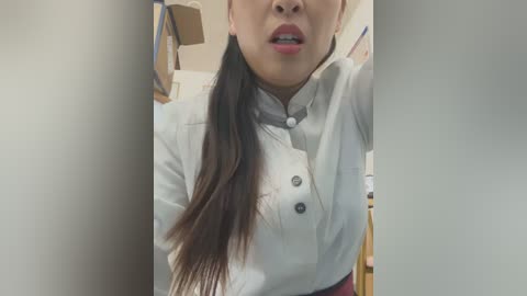 Media: A video of an Asian woman with long black hair in a ponytail, wearing a white chef's jacket with buttons, and a red apron, standing in a dimly lit kitchen with cardboard boxes in the background.