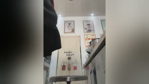 Video of a modern caf\u00e9 with white walls, minimalist decor, and framed drink menus. A black jacket hangs on the left, and a wooden counter holds a coffee maker and cups on the right.