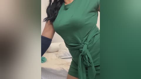 Video of a woman with medium brown skin wearing a green, fitted, short-sleeved dress tied at the waist, standing in a bedroom with white bedding and a pillow.