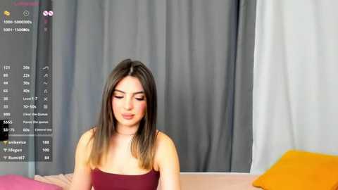 Media: Video of a young woman with straight brown hair, wearing a strapless maroon top, seated on a bed with a pink and orange blanket. Gray curtains in the background.