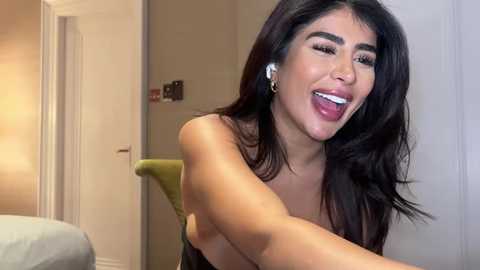 Media: A video of a smiling, topless Latina woman with long, straight black hair, wearing white earbuds, in a modern bedroom with beige walls and a green chair.