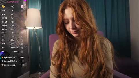 Media: A video of a fair-skinned, red-haired woman with long, wavy hair, wearing a beige sweater, sitting in a dimly lit room with teal curtains and a lit lamp.