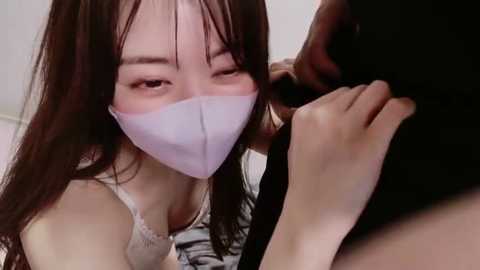 A video of an Asian woman with long black hair and fair skin, wearing a white surgical mask, engaging in a close-up oral sex act with a dark-skinned man.