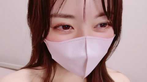 Video of an East Asian woman with straight brown hair, wearing a white face mask, focusing on her eyes and nose, with a plain white background.