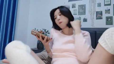 Media: Video of an East Asian woman with short black hair, wearing a light pink sweater, seated on a bed, holding a phone, in a bedroom with blue curtains, white wall, and hanging photos.