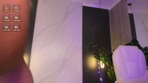 Media: Video of a modern living room with sleek white marble walls, a dark wooden cabinet, a white chair, and a potted plant under soft purple lighting.