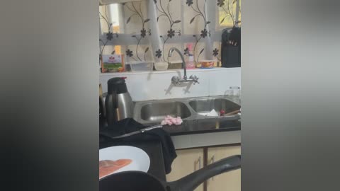 A video of a small kitchen sink with a stainless steel faucet, black countertop, and a black kettle. The background features a white cabinet with floral wallpaper and a microwave.