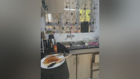 Video of a modern, cluttered kitchen with white cabinets, black countertops, and a stainless steel sink. Toasted bread, an orange juice bottle, and a black kettle sit on a plate, while a pink dishcloth lies on the counter.