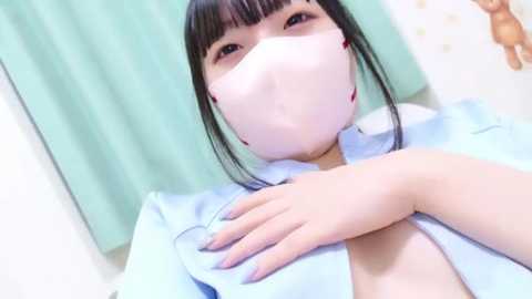 Video of an East Asian woman with straight black hair and a light blue surgical mask, partially lifting her hospital gown to reveal a cleavage. Background features a green curtain and a white wall.