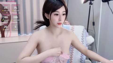 Media: A video of a young, slender, East Asian woman with light skin, dark hair, and small breasts, topless, sitting in a modern room with pastel decor.