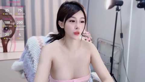 Media: Video of an East Asian woman with fair skin, long dark hair, and pink lipstick, wearing a strapless pink dress, in a modern, softly lit bedroom with a soft bed and a standing lamp.