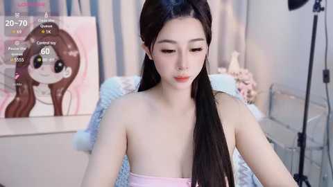 Media: Video of an East Asian woman with long black hair, light skin, and a slender physique, wearing a pink strapless top, sitting in a softly lit room with a colorful anime painting on the wall.