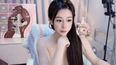 Media: Video of a young, fair-skinned Asian woman with long, straight black hair, sitting topless, in a softly lit room. She has a serene expression. Background includes a pink poster, fluffy blanket, and camera equipment.