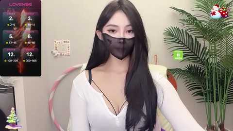 Media: A video of an Asian woman with long black hair, wearing a white top and a black face mask, sitting in a room with a plant and a pink hula hoop.