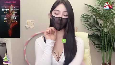 Media: A video of an East Asian woman with long black hair and light skin, wearing a white top and a black mask, sitting indoors with a green plant in the background.