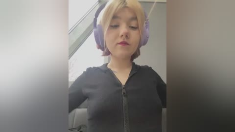 Media: Video of a young woman with short, platinum blonde hair wearing large purple headphones and a black zip-up hoodie. She has fair skin and a neutral expression.