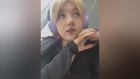 A video of a young woman with light skin, blonde hair, and purple headphones, wearing a black hoodie, looking up with a contemplative expression.