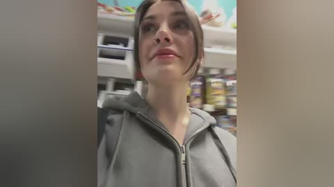 Video of a young woman with light skin and brown hair in a ponytail, wearing a gray hoodie, standing in a brightly lit grocery store aisle with shelves of various products in the background.