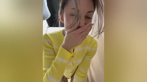 Media: Video of a young Asian woman with light skin, wearing a yellow-striped sweater, covering her face with both hands, in a dimly lit room.