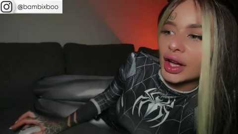 Media: Video of a young woman with fair skin, long black and green hair, wearing a black Spider-Man shirt, lying on a dark sofa.