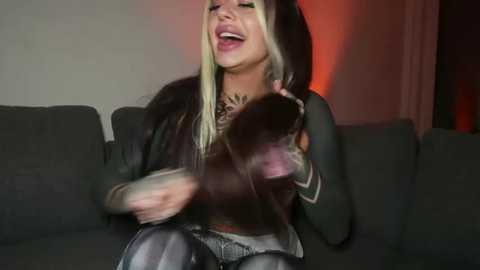 Media: Video of a laughing, blonde woman with long hair, wearing a black hoodie and jeans, holding a small, brown dog on a dark gray couch under dim lighting.