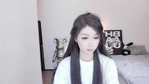 Media: Video of a young woman with long, straight black hair, wearing a white shirt, sitting in a minimalist bedroom with beige walls, a bed, and a stuffed animal.