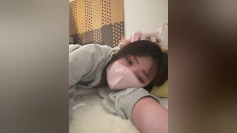 A video shows an Asian woman with brown hair and light skin lying on a bed, wearing a gray hoodie and face mask, looking tired. The background features a wall with patterned brown and yellow wallpaper.