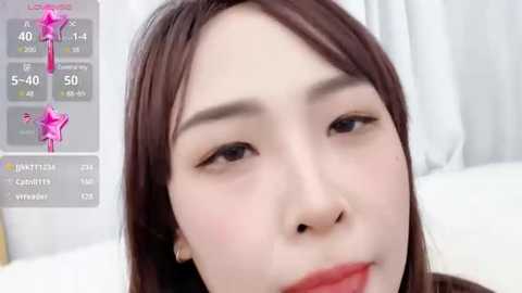 Media: A close-up video of an East Asian woman with fair skin and long, straight dark brown hair. She has a neutral expression and is wearing light makeup. The background features a white bedsheet.