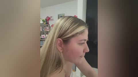 Media: Video of a young woman with long blonde hair in a side profile, sitting on a bed, in a softly lit bedroom with shelves and red decorations in the background.