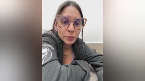 Media: Video of a Latina woman with medium skin tone, dark hair, wearing large pink-tinted glasses and a dark hoodie, sitting indoors with a neutral background.