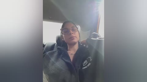 Media: A video of a young woman with light brown skin and dark hair, wearing glasses and a dark jacket, taking a selfie in a car.