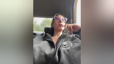 Video of a woman with dark hair, wearing sunglasses and a gray hoodie with a logo, seated in a car, with blurred background and sunlight.