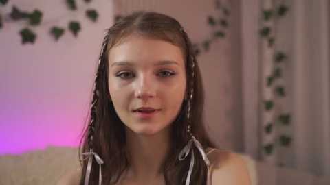 A video of a young woman with straight, dark brown hair styled in two braids, wearing a white tank top, standing against a softly lit background with hanging green ivy and a purple light.