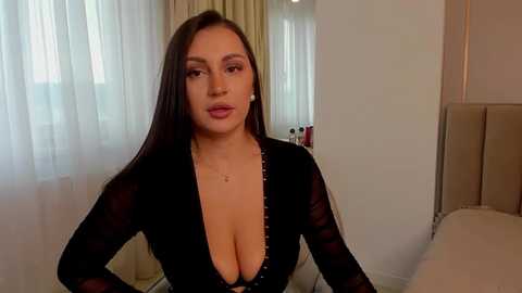 Media: Video of a curvy woman with long dark hair and fair skin, wearing a plunging black lace bodysuit, standing indoors in a modern, well-lit room with white curtains and beige furniture.