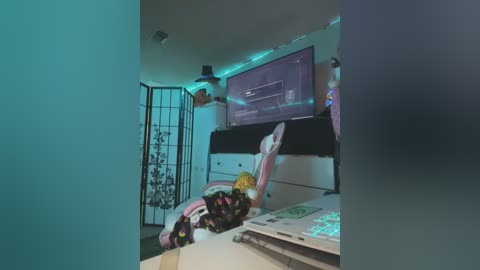 A video of a dimly lit, futuristic gaming room with a pink and black gaming chair, monitor displaying code, neon-lit LED strip, and a pink plush toy on the floor.