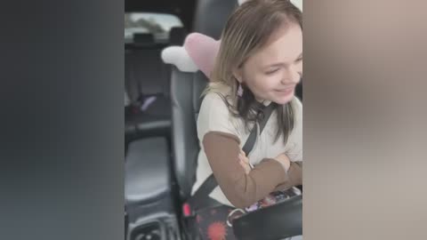 Media: Video of a young girl with blonde hair and a pink bunny headband, smiling, sitting in a car seat, wearing a brown sweater and black pants.