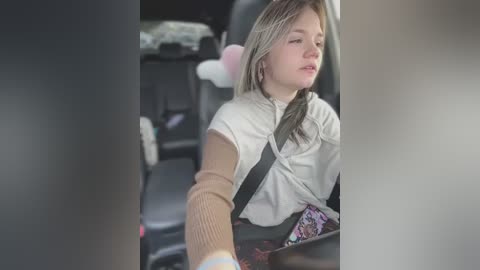 Media: Video of a young Caucasian woman with shoulder-length blonde hair, wearing a beige sweater and black shoulder harness, seated in a car, appearing tired and possibly asleep.