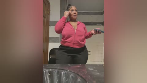 Video of a plus-size Black woman with a curvy figure in a pink zip-up hoodie and black leggings, applying makeup in a small bathroom with a sink, white tiles, and a wooden door.