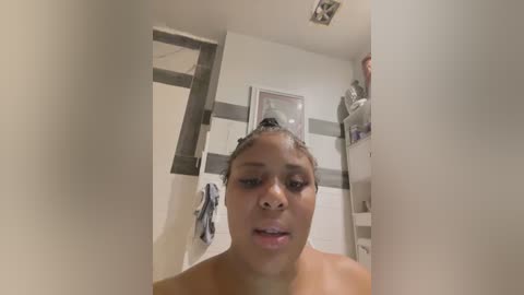 Media: Video of a young African-American woman with medium brown skin, dark curly hair in a bun, and full lips. She is topless in a modern bathroom with white walls, grey tiles, and a towel hanging on a hook.