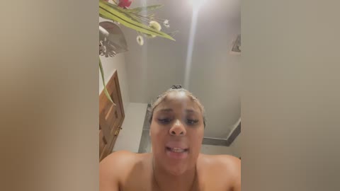 Video of a nude Black woman with light skin tone, dark curly hair, and small breasts, standing in a modern bathroom with beige walls, floral decorations, and a mirror reflecting her upper body.